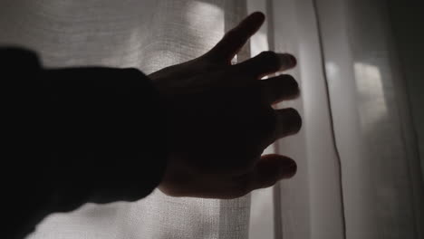 Hand-reaching-towards-white-window-curtain---opening-fabric-to-let-sun-shine-inside-dark-room