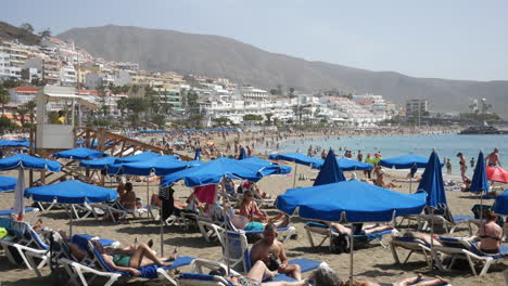 Bustling-beach-scene-by-the-ocean-with-under-blue-parasols,-with-a-lively-backdrop-of-a-coastal-town-of-Los-Christianos-in-Tenerife-with-mountains