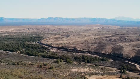 Long-Freight-Train-Carrying-Cargo-Through-The-Desert-Mountains,-Border-Between-California-and-Arizona-,-Colorado-River-,-I-40-freeway-East,-Wide-Angle-CInematic-Aerial-Establishing-Shot