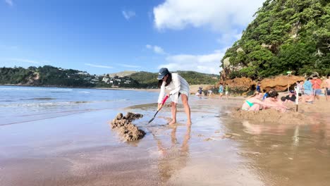 A-young-Asian-woman-in-a-white-shirt-digs-with-a-shovel-on-the-beach-near-a-group-of-children-on-a-bright-sunny-day