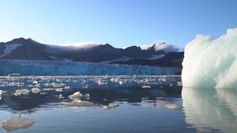 Iceberg-and-Pieces-of-Ice-in-Glacial-Lagoon-Under-Glacier-and-Mountain-Hills-of-Svalbard,-Norway