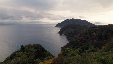 Aerial-drone-above-red-green-orange-trees,-Kyotango-Kyoto-Japanese-sea-mountain-landscape-in-cloudy-daylight,-sunshine-through