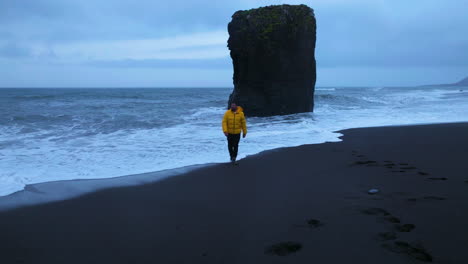 Front-Portrait-Of-Male-Traveler-In-Yellow-Jacket-Strolling-On-Black-Sand-Beach-Laekjavik-In-Iceland