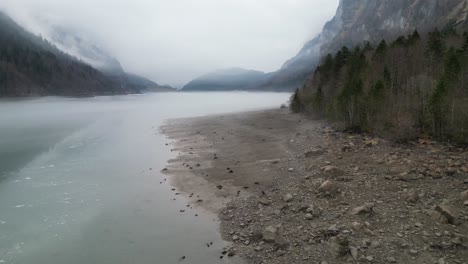 Drone-Vuela-A-Lo-Largo-De-La-Costa-Gris-Congelada-Del-Lago-Embalse-Con-Nubes-Brumosas-Que-Rodean-Las-Montañas-En-La-Distancia