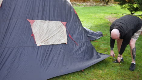 Un-Hombre-Derribando-Una-Tienda-De-Campaña-Quitando-Clavijas-Bajo-La-Lluvia-En-Un-Campamento-Mientras-Acampaba.