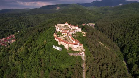 Rasnov-citadel-amidst-green-forests-under-a-clear-sky,-daylight,-aerial-view