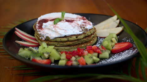 Slow-motion-pullback-shot-of-plate-with-healthy-green-oat-and-matcha-pancakes-served-with-apple-kiwi-and-strawberry-slices-and-whipped-cream