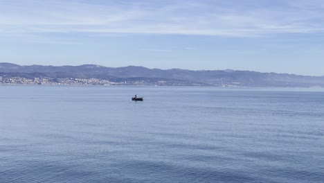 Fisherman-in-a-boat-on-Sea-shore-with-town-in-distance,-Opatija,-Croatia