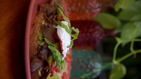 Vertical-Slow-motion-dolly-in-shot-of-a-beef-deli-sandwich-topped-with-arugula-mozzarella-cheese-pickles-cucumber-and-tomato-served-with-salad