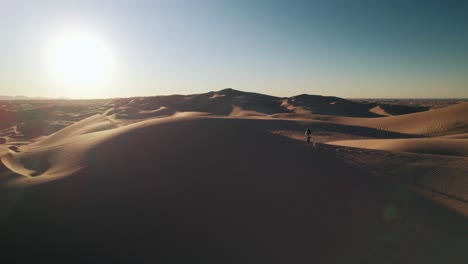 Ein-Mann-Läuft-Bei-Sonnenaufgang-In-Der-Glamis-Sand-Dunes-Wüste-Zwischen-Unendlichen-Dünen,-Während-Eine-Drohne-Ihn-Aufzeichnet,-Es-Ist-Goldene-Stunde-Und-Ein-Klarer-Blauer-Himmel