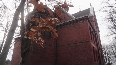Large,-red-brick-building-with-intricate-architectural-designs,-surrounded-by-bare-trees-and-fluttering-leaves-under-an-overcast-sky,-creating-a-somber-atmosphere