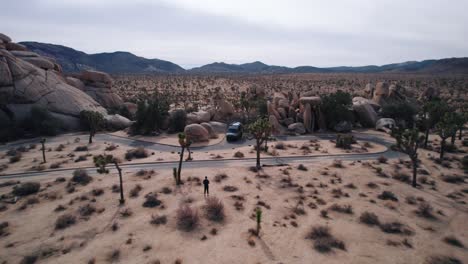 Ein-Mercedes-Sprinter-Van-Parkt-Im-Joshua-Tree-Nationalpark,-Während-Eine-Drohne-Rückwärts-Von-Ihm-Wegfliegt.-Im-Video-Sind-Schroffe-Felsformationen-Und-Joshua-Trees-Zu-Sehen.