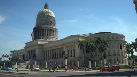 Capturing-the-Capitolio-building-being-repaired-in-2018,-embraced-by-pine-trees,-offering-a-nice-view-of-the-clouds,-amidst-the-bustling-scene-of-vintage-1950s-cars-and-the-waving-Cuban-flag