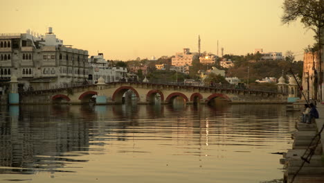 Con-El-Puente-Chandpole-En-El-Lago-Pichola-En-Udaipur,-India,-Con-Un-Fascinante-Telón-De-Fondo-De-Hermosa-Arquitectura-Blanca-Al-Fondo-Justo-Después-Del-Atardecer