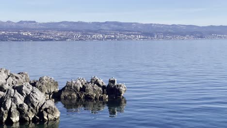 Seagulls-resting-on-boulders-on-Sea-shore-with-town-in-distance,-Opatija,-Croatia
