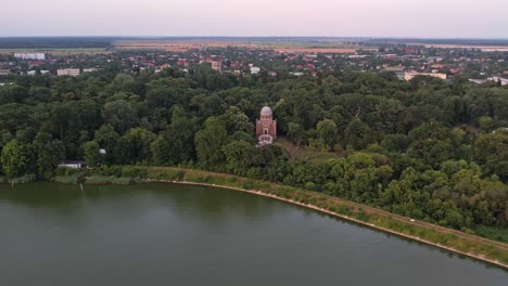 Una-Iglesia-Histórica-Rodeada-De-Vegetación-Cerca-De-Un-Lago-Al-Amanecer,-Vista-Aérea