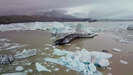 Fliegen-Sie-Zwischen-Zwei-Eisberggipfeln,-Die-In-Einer-Schlammigen-Gletscherlagune-In-Island-Schwimmen