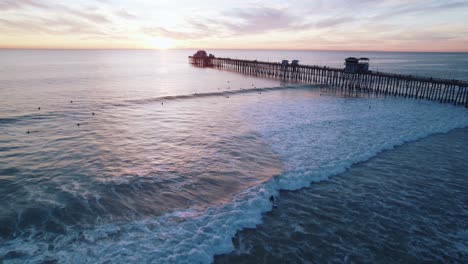 Aerial-drone-view-of-surfers-riding-gentle-waves-near-a-pier-during-a-beautiful-sunset,-capturing-the-essence-of-tranquility-and-leisure