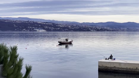 Pescador-En-Barco-Estacionado-En-La-Orilla-Del-Mar-Con-La-Ciudad-A-Distancia-Cerca,-Opatija,-Croacia