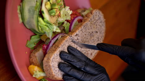 Vertical-slow-motion-of-a-person-wearing-black-latex-gloves-cutting-in-half-a-whole-wheat-bread-with-visible-onions-and-pickles