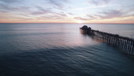 Eine-Ruhige-Meereslandschaft-Bei-Sonnenuntergang-Am-Kalifornischen-Meer-Mit-Einem-Sich-Abzeichnenden-Pier-Am-Meer-Und-Surfern,-Die-Wellen-Fangen,-Aufgenommen-Mit-Einer-Drohnen-Luftaufnahme