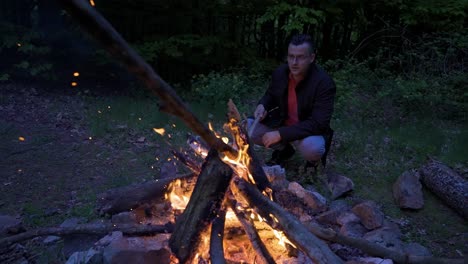 Sitting-by-the-bonfire,-the-camper-is-adding-more-wood-to-the-fire-to-keep-the-flames-glowing,-at-a-campsite-on-Strandzha-Mountains-in-Bulgaria