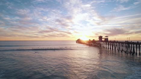 Eine-Ruhige-Meereslandschaft-Bei-Sonnenuntergang-Am-Kalifornischen-Meer-Mit-Einem-Sich-Abzeichnenden-Pier-Am-Meer-Und-Surfern,-Die-Wellen-Fangen,-Aufgenommen-Mit-Einer-Drohnen-Luftaufnahme