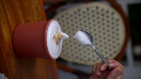 Vertikale-Zeitlupe-Einer-Weiblichen-Hand,-Die-Mit-Einem-Metalllöffel-Mit-Dem-Schaum-Eines-Cappuccinos-Spielt