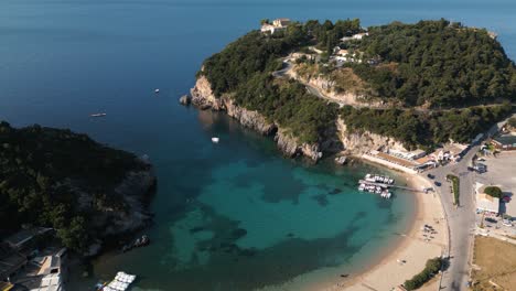 Aerial-estabilshing-orbit-above-golden-sandy-beach-and-cove-in-Corfu-Greece-with-boats-at-dock-and-tourists-on-beach