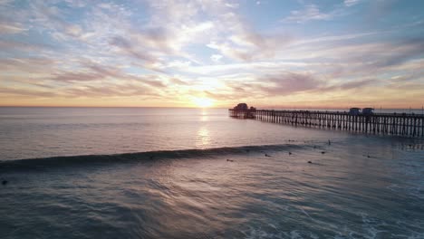 Eine-Ruhige-Meereslandschaft-Bei-Sonnenuntergang-Am-Kalifornischen-Meer-Mit-Einem-Sich-Abzeichnenden-Pier-Am-Meer-Und-Surfern,-Die-Wellen-Fangen,-Aufgenommen-Mit-Einer-Drohnen-Luftaufnahme