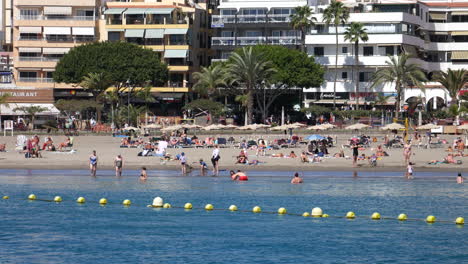 Strand-Von-Los-Christianos,-Teneriffa-–-Sonnige-Strandszene-Mit-Menschen-Zum-Sonnenbaden-Und-Schwimmen,-Palmen-Und-Gebäuden-Am-Strand