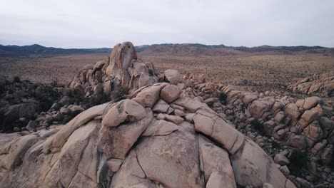 Drones-Volando-En-El-Parque-Nacional-Joshua-Tree-Sobre-Formaciones-Rocosas-Escarpadas-Mientras-Un-Hombre-Solitario-Se-Encuentra-En-La-Cima-De-La-Roca