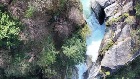 A-vibrant-turquoise-waterfall-carving-through-rocky-terrain-surrounded-by-lush-greenery,-aerial-view