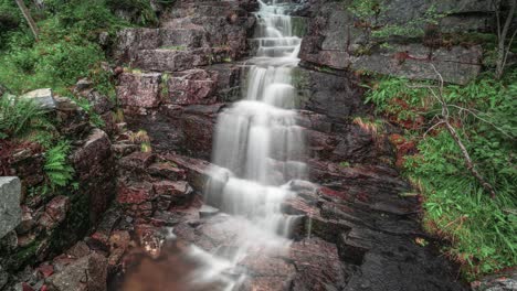 A-timelapse-video-of-a-small-waterfall-cascading-over-the-dark-rocks-in-the-summer-forest