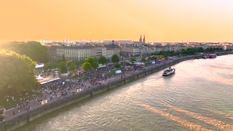 Garonne-River-shore-during-Wine-Fair-full-of-crowds-at-sundown,-Aerial-approach-shot