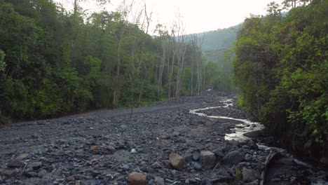 A-low-drone-shot-tracking-a-rocky-dry-river-which-is-surrounded-by-forest-trees-during-sunrise-in-Risaralda,-Colombia