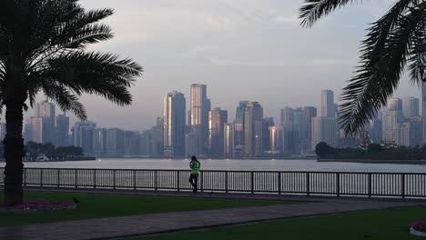 Un-Residente-Disfrutando-De-Un-Paseo-Matinal-Por-La-Corniche-De-Sharjah-Con-La-Vista-Del-Horizonte-De-Sharjah-Al-Fondo