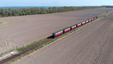 Aerial:-steam-narrow-gauge-railway-in-the-countryside-passing-by-sown-fields-and-with-the-sea-in-the-background