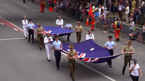 Soldados-De-Las-Tres-Ramas-De-La-Fuerza-De-Defensa-Australiana,-La-Marina-Real-Australiana,-El-Ejército-Australiano-Y-La-Fuerza-Aérea-Real-Australiana-Sosteniendo-Las-Banderas-Marchando-Por-La-Calle-En-El-Desfile-Del-Día-De-Anzac