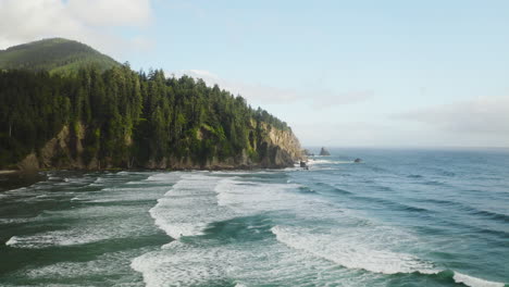 Sunlight-hitting-forested-Oregon-Coast-hillside,-aerial-over-Pacific-Ocean-waves