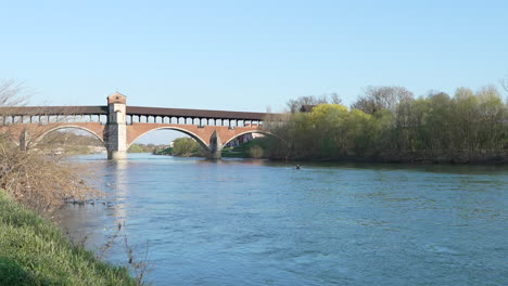 Nice-view-of-Ponte-Coperto-is-a-bridge-over-the-Ticino-river-in-Pavia-at-sunny-day,-man-sailing-on-a-canoe,-Lombardy,-Pavia,-Italy