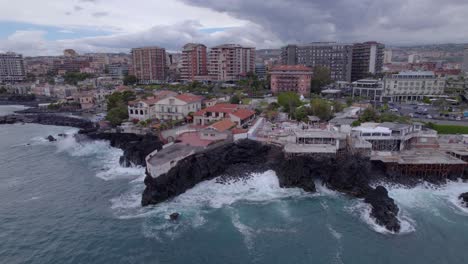 Aerial-trucking-left-shot-of-the-waterfront-of-Catania,-Sicily,-Italy-with-sea-waves-crashing-on-the-volcanic-cliff