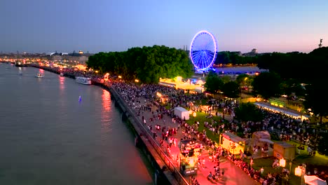 Ferris-wheel-in-neon-lights-and-crowds-in-Bordeaux-France-at-sundown,-Aerial-flyover-shot