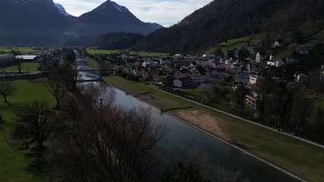 Drone-clip-over-calm-river-passing-traditional-Alpine-village-in-Switzerland,-surrounded-by-mountains