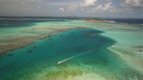Kitesurfistas-Deslizándose-Sobre-Un-Mar-Turquesa-Claro-Con-Un-Arrecife-De-Coral,-Vista-Aérea