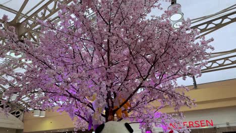 Artificial-cherry-blossom-tree-in-a-busy-mall-interior,-daylight-coming-through-the-skylight,-shoppers-walking
