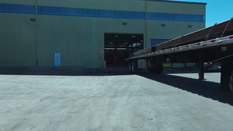 Low-angle-dolly-follows-semi-truck-with-steel-beams-reversing-into-warehouse-at-midday