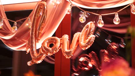 Das-Wort-„Liebe“-Aus-Rosa-Luftballons,-Mit-Einer-Glühbirnengirlande-Im-Hintergrund,-Panorama