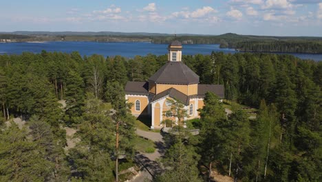 Laukaa-Church,-Finland,-orbiting-drone-shot-of-the-old-wooden-church-amidst-lakes-and-forest-on-a-beautiful-summer-day