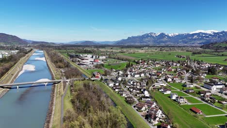 Ruggell-in-liechtenstein-with-river,-village,-and-snow-capped-mountains-in-the-background,-on-a-clear-day,-aerial-view